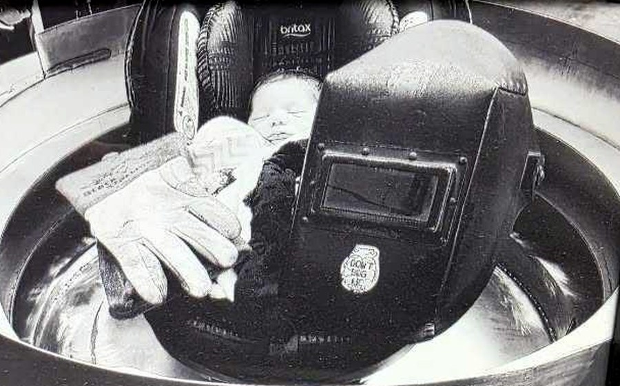 Sean's newborn daughter Annie asleep in a car seat set inside a stainless feeder bowl, beside a welding helmet and gloves