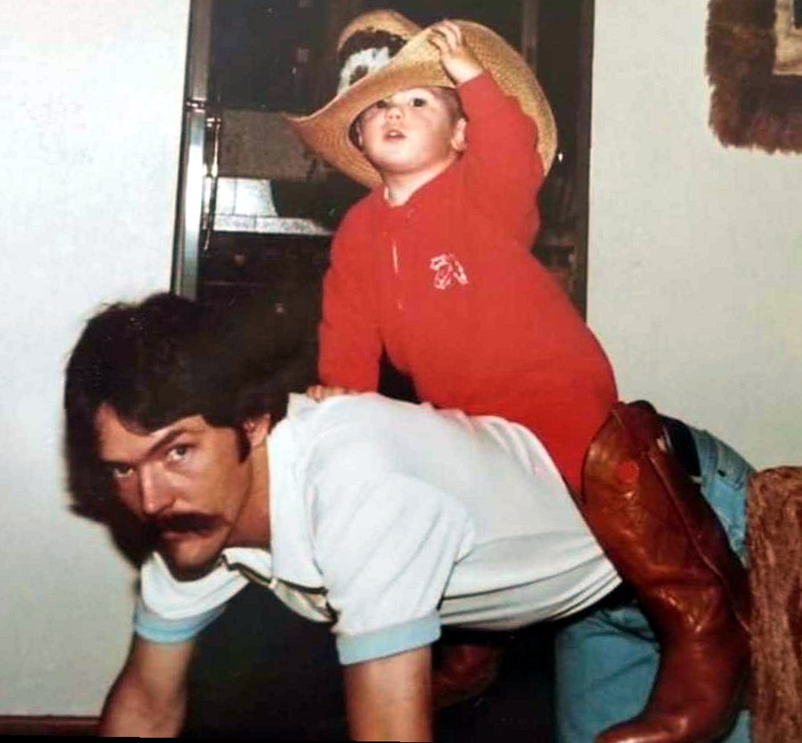 Doug Wilcox on the floor with his young son Sean riding on his back in a straw cowboy hat